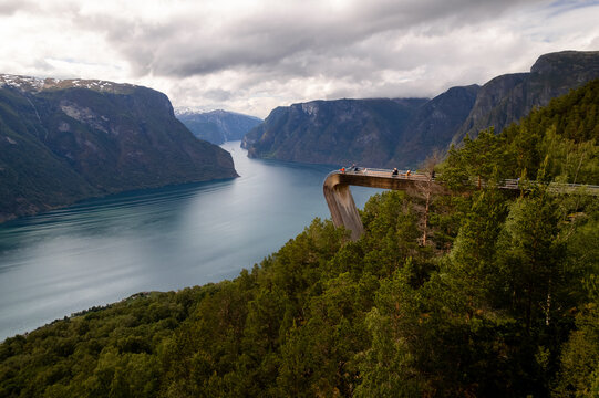 Aerial view of the Stegastein viewpoint jutting out over the Aurlandsfjord, with deep blue waters contrasting against the lush green forests, Aurland, Vestland, Norway.