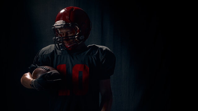 American football player holding ball with quiet reflective emotion under spotlight. Concept of personal mindset training, tutorials, introspective sport content, branding.