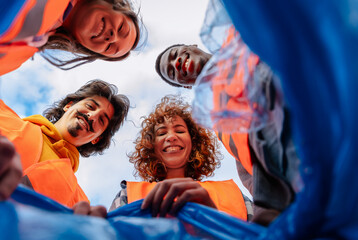 Smiling group of multiracial volunteers in vests look down while putting trash in a blue recycling bag.