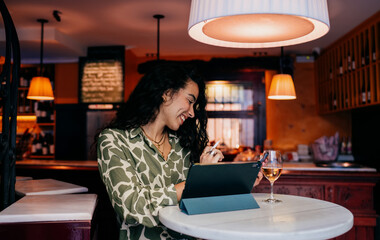 Laughing woman writes on digital tablet, glancing at smartphone, expressing joy and ease in mobile creative workflow, surrounded by warm light and modern freelance identity.