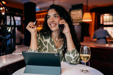 Smiling woman talks on smartphone while holding stylus, tablet open on bar table, radiating positive energy and confidence in cozy workspace filled with warm light and wine.