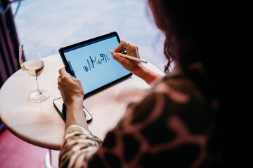 Freelancer sketches digitally on tablet with stylus in bright cafe, focusing on fluid lettering while surrounded by light, tech, and wine, merging creativity and mobile work freedom.