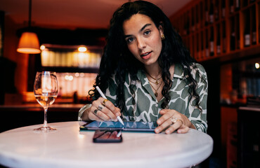 Freelancer works on digital sketch using stylus and tablet, wine beside, capturing quiet focus and tactile interaction with mobile creative tools in warm-lit workspace.