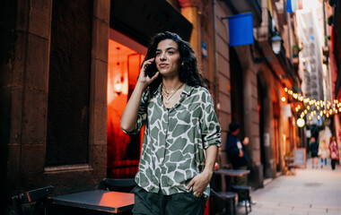 Calm woman with phone walks slowly past street café, speaking on call with thoughtful expression, reflecting presence, communication and independence in mobile lifestyle.