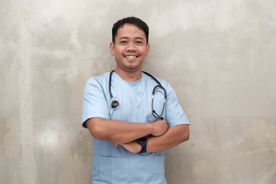 Happy Southeast Asian male nurse in scrubs with stethoscope, arms crossed, isolated on wall, ideal for healthcare ads and medical content.

