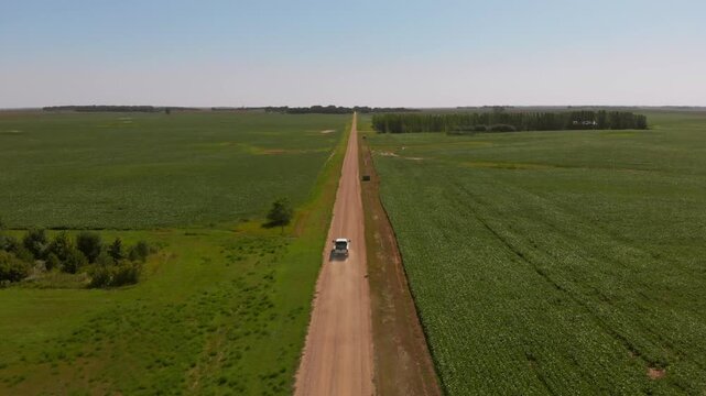 Aerial shot slowly tracking a white pickup truck driving down a long dirt road between wide green fields and scattered wetlands.