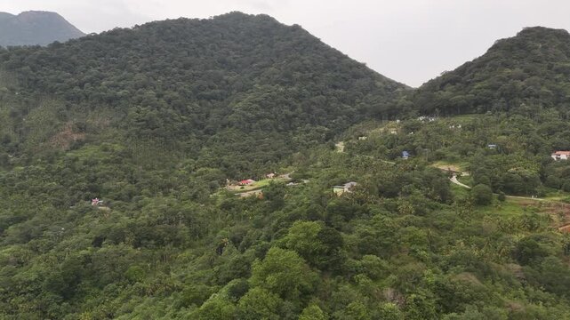 A wide aerial shot of a densely forested mountain range. Small, colorful houses are scattered across the hillside, connected by a winding road, showing a remote settlement nestled in nature.