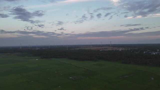A wide aerial landscape view captured near dawn, showing a field of modern wind turbines stretching across the horizon. The foreground features lush green agricultural fields and trees.