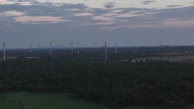 Scenic aerial footage showing wind turbines on hills, a lake with rocky terrain, and a vast mountain range under moody twilight clouds. Drone Footage of Wind Turbines Overlooking a Lake and Mountains