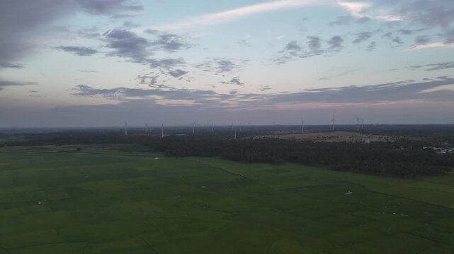 Aerial view of nature and technology. A tranquil lake reflects the moody evening sky, set within a vast green valley. On the surrounding hills and plains, dozens of wind turbines stand tall