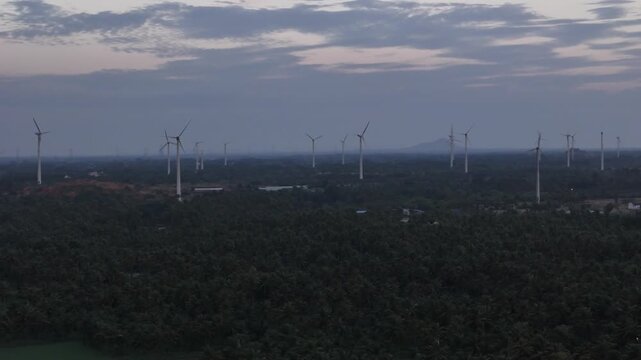 Wide aerial footage near dusk showing a field of modern wind turbines stretching across the horizon. The foreground features lush green agricultural fields and tree. renewable energy generation