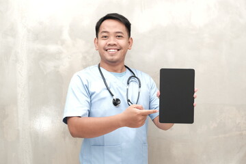 Smiling Southeast Asian male nurse or doctor in scrubs pointing at blank tablet screen, ideal for health advertisements and app promotions.
