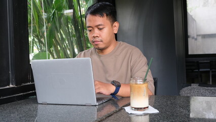 Focused Southeast Asian man using laptop in modern cafe with iced coffee, perfect for freelance...