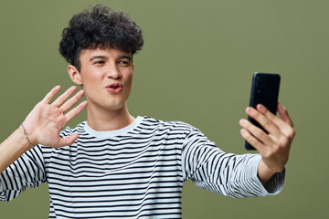 young man taking selfie with smartphone, wearing striped shirt, making playful gesture, green background, casual style, indoors