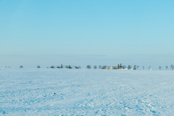 Expansive winter landscape featuring a serene snowy field under a clear blue sky, with distant buildings and trees creating a tranquil rural scene with copy space