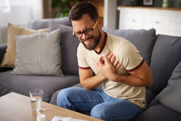 Obraz premium A young man sits on a sofa at home, looking worried while holding his chest. He appears to be feeling chest pain, with a glass of water on the table nearby.