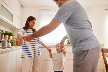 A joyful family enjoys time together in a warm kitchen. Parents hold hands with their young child as they dance and laugh in happiness during the afternoon.