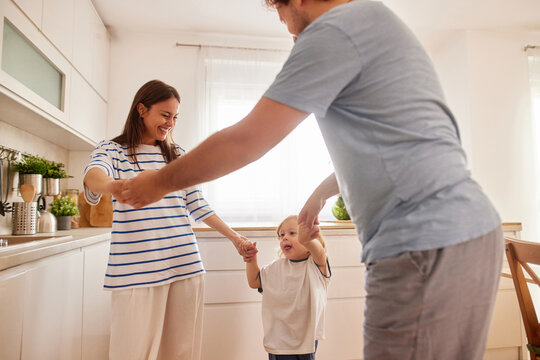 A joyful family enjoys time together in a warm kitchen. Parents hold hands with their young child as they dance and laugh in happiness during the afternoon.