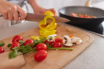 A person is chopping bright yellow bell peppers, cherry tomatoes, and mushrooms on a wooden cutting board while cooking in a warm kitchen atmosphere.