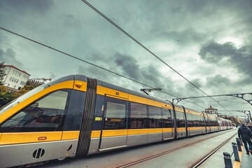 Fototapeta premium A yellow tram rolls along its track in a bustling city, blending modern transportation with stunning architecture and dark, dramatic clouds hovering overhead, hinting at an approaching storm