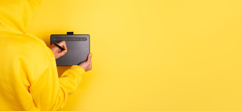 Person in yellow hoodie drawing on a graphic tablet with stylus against solid yellow background, creative workspace concept. - Powered by Adobe