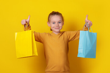 Fototapeta premium Smiling young girl in hoodie holding colorful shopping bags and showing thumbs up against bright yellow background.
