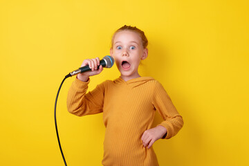 Excited young girl in yellow holding a microphone, singing with a surprised expression against a bright yellow background.
