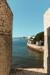 A beautiful waterfront scene is framed by a weathered stone archway. Calm waters reflect the clear blue sky while lush greenery lines the shore, creating an inviting atmosphere