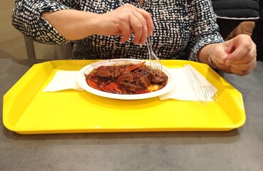 Upper body and hands of a woman eating Chinese food from a yellow tray with a plastic plate and plastic fork.