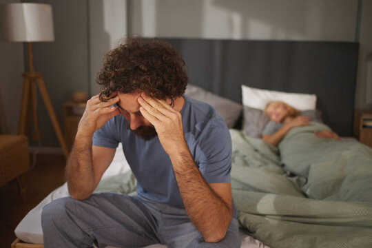 A man sits on the edge of a bed, holding his head in frustration, while a woman lies under the covers, emotionally distant. Tension fills the room. - Powered by Adobe