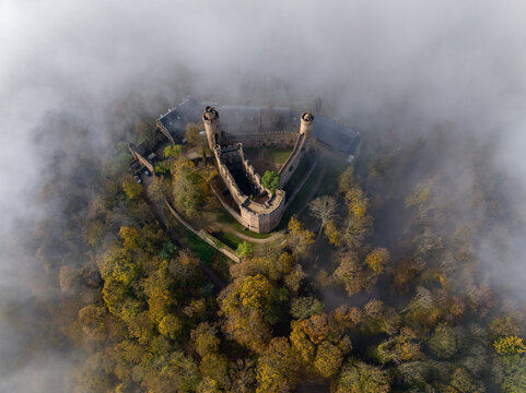 Aerial view of the stark, stone Auerbach Castle ruins piercing through a swirling sea of ethereal fog, perched atop autumn-kissed trees, Bensheim, Hessen, Germany.