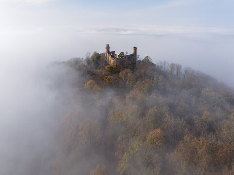 Aerial view of a medieval castle Auerbach perched atop a hill, veiled in ethereal mist, with the surrounding forest displaying autumn hues, Bensheim, Hessen, Germany.