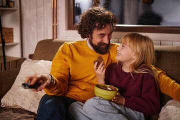 A smiling man and a young girl share a joyful moment while watching a movie on the couch. They enjoy snacks and laughter in a warm and inviting living room.