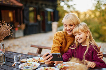 A mother and daughter share a joyful moment in an autumn setting, smiling and using a smartphone to take a selfie while surrounded by tasty dishes.