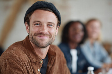 Fototapeta premium Confident, friendly man smiles at camera. Diverse group blurred in background. Represents leadership, teamwork, creativity, community. Use for business, education.