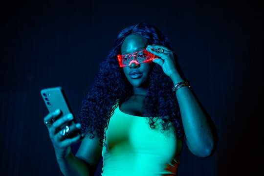 Woman wearing illuminated glasses and holding a phone in a dark, neon lit environment