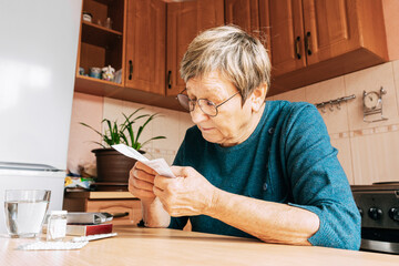 An elderly woman reads pill instructions in kitchen setting. Health maintenance, healthy aging, vitamin support, home treatment