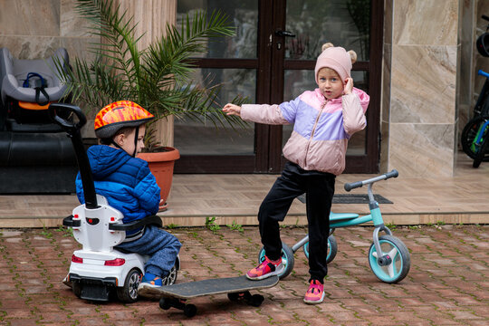 Two young siblings playing with a tricycle, ride-on car, and skateboard outdoors, showing fun, interaction, and childhood activity