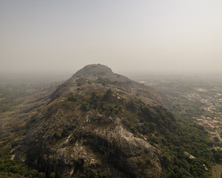 Aerial view of the rugged, rocky landscape of Mount Patti, its peak shrouded in mist, contrasting with the green trees dotting its slopes, Ado Awaye, Oyo, Nigeria..