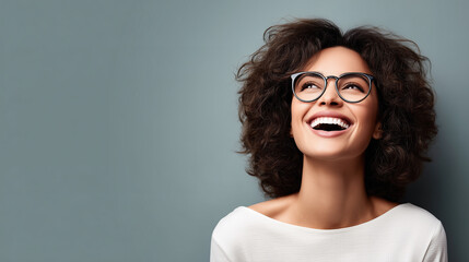 Joyful young woman with curly hair and stylish glasses smiles brightly against a simple blue background with copy space.