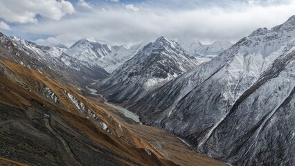 Expansive view of snow-covered mountain peaks surrounding a winding river in a valley, with rocky terrain and patches of brown grass, creating a serene and dramatic landscape atmosphere