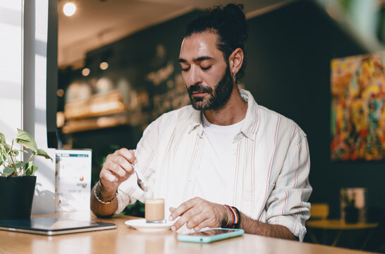 Man stirring coffee at cafe table with smartphone beside, focused moment, casual attire, relaxed remote worker enjoying balance of technology and personal lifestyle.