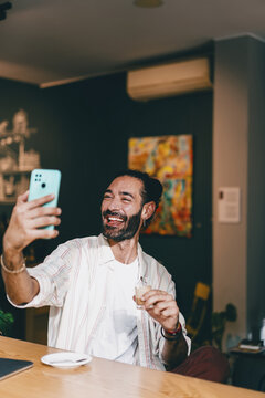 Smiling man raises smartphone to snap selfie, enjoying espresso at stylish caf&eacute;, embracing personal freedom and identity through mobile tech and digital self-representation.