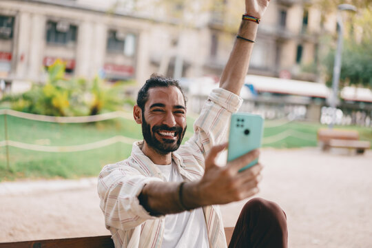 Excited man smiles widely during video call, arm raised mid-gesture, sharing enthusiastic reaction in personal mobile space with tech-driven connection and vibrant energy.