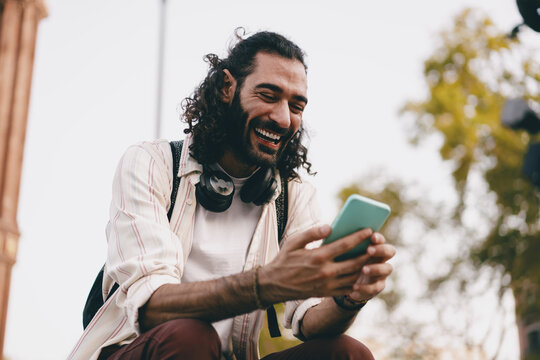Smiling man sits with smartphone outdoors, laughing while looking at screen, enjoying casual digital interaction and mobile lifestyle.