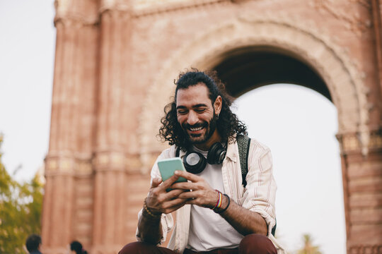 Man with curly hair smiles at smartphone while seated outdoors, showing joy, digital connection and casual freedom in modern tech-driven lifestyle.