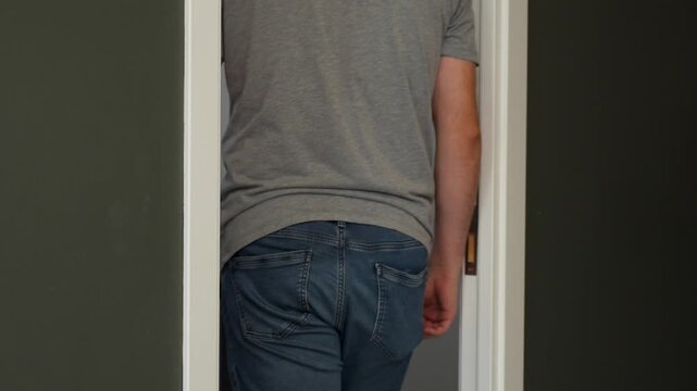 Back view of a man wearing a grey T-shirt and jeans as he opens a white door and walks into a room