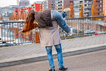 Young happy couple walking playfully canal in modern European city, man giving woman piggyback ride city canal bank, Playful autumn date, partnership and support, Urban romance, modern architecture