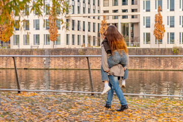 Young happy couple walking playfully canal in modern European city, man giving woman piggyback ride city canal bank, Playful autumn date, partnership and support, Urban romance, modern architecture