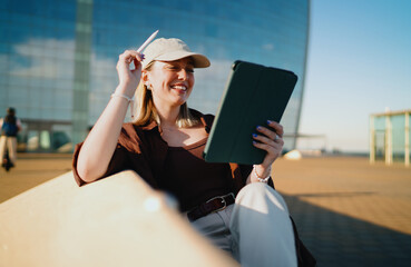 Young woman in cap smiling while working on tablet outdoors, using stylus with focus, symbolizing freelance independence, creativity, mobile technology and digital lifestyle. © BullRun
